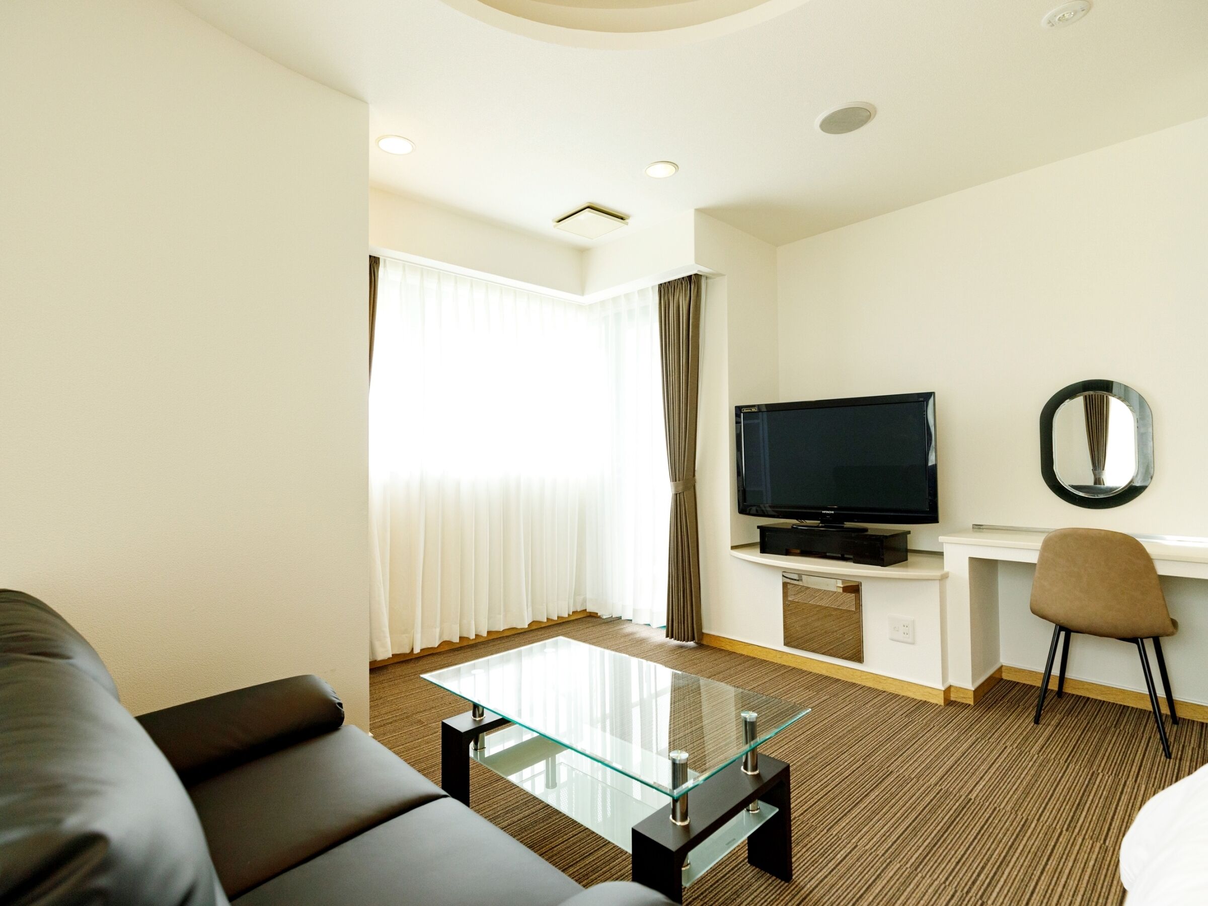 Una camera d'albergo moderna con un tavolino in vetro, un divano nero, una TV a schermo piatto su un mobile bianco, una piccola scrivania con una sedia e un'ampia finestra con tende.