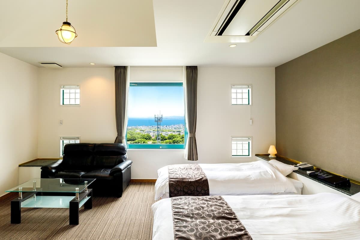 A hotel room with two beds, a sofa, a glass coffee table, and a view of the city and sea.