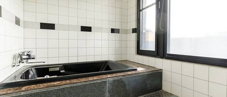 A modern bathroom with a black bathtub, white tiled walls, and a window with blinds.
