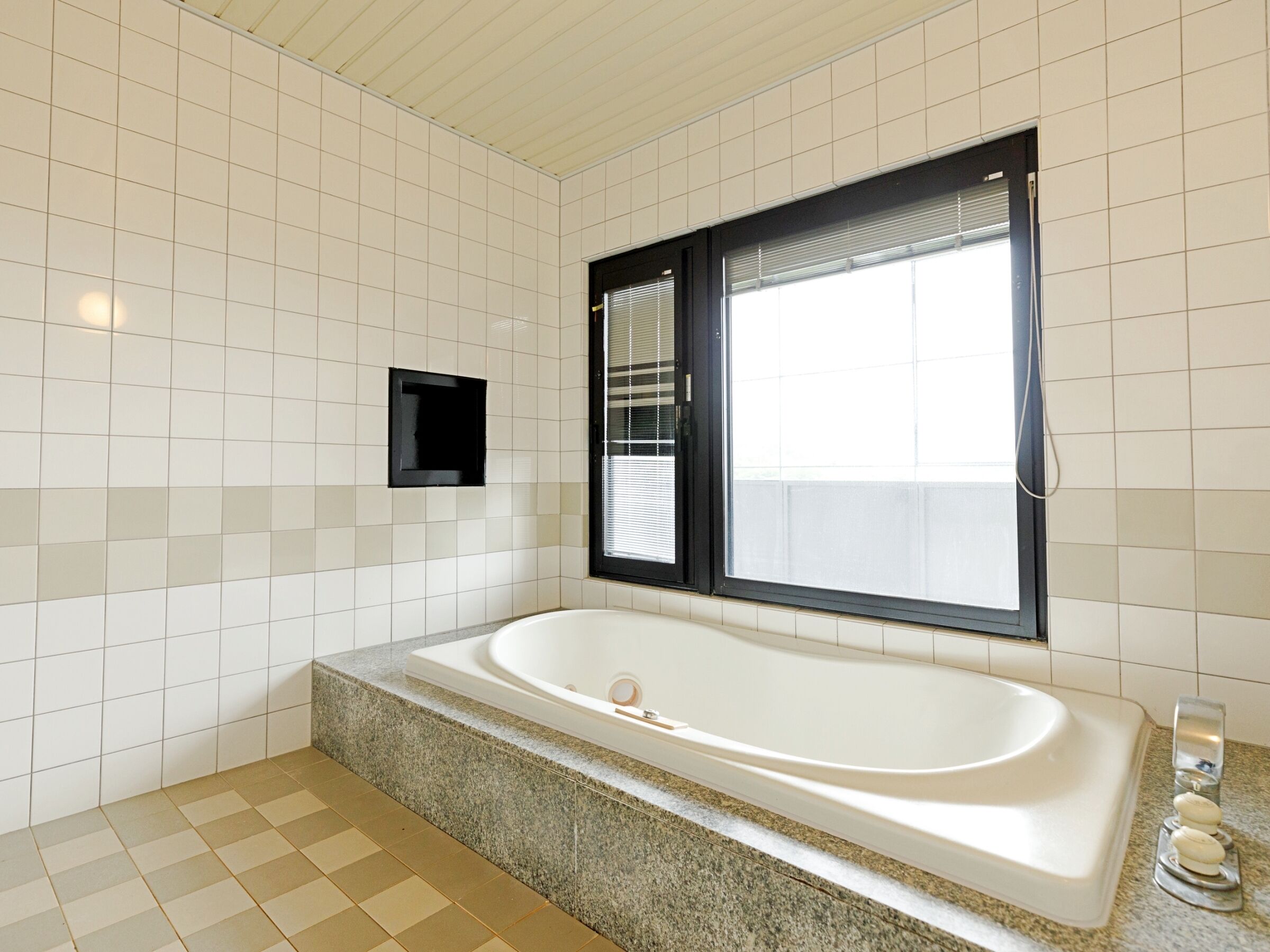 A modern bathroom with a freestanding bathtub, a large window with blinds, and a small TV mounted on the wall.