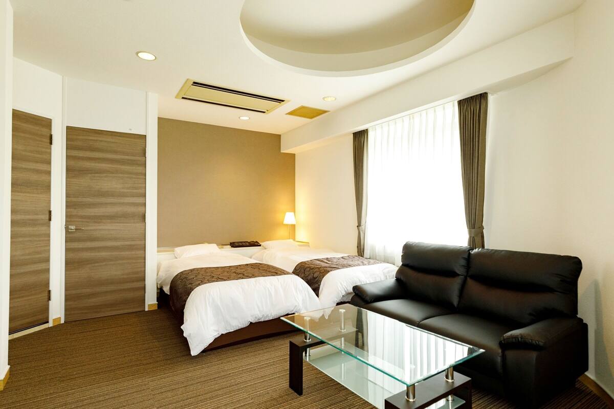 A hotel room with a bed, a glass coffee table, a leather sofa, and a window with curtains.