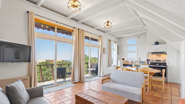 A spacious living area with a sofa, a wooden coffee table, and a dining table with chairs. There is a kitchen area with a stove and oven, and a balcony with a view of the greenery.