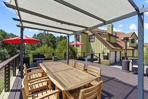 Una terrazza esterna con tavolo e sedie in legno, due ombrelloni rossi e un barbecue.