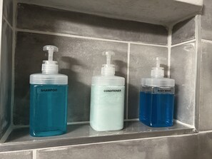 Drei Flaschen Flüssigkeit auf einem Regal: Shampoo, Conditioner und Duschgel.