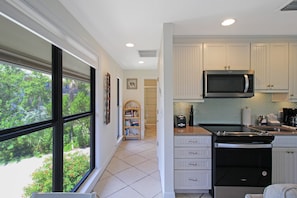 A kitchen with white cabinets, a microwave, a toaster, and a stove. There is a window with a view of greenery outside. - Enjoy pool views from your Sanibel Island Villa - private beach - pet friendly - Signal Inn 04 (Sanibel)