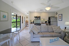 A modern kitchen with white cabinets, a refrigerator, and a microwave. There is a dining area with a table and chairs, and a sofa in the living room. - Enjoy pool views from your Sanibel Island Villa - private beach - pet friendly - Signal Inn 04 (Sanibel)