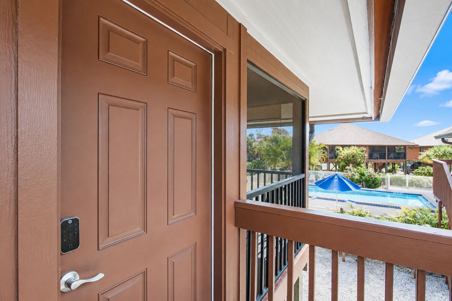 Une porte marron dotée d’un clavier de code et d’une poignée, qui donne sur un balcon offrant une vue sur une piscine et un bâtiment au toit en pente.