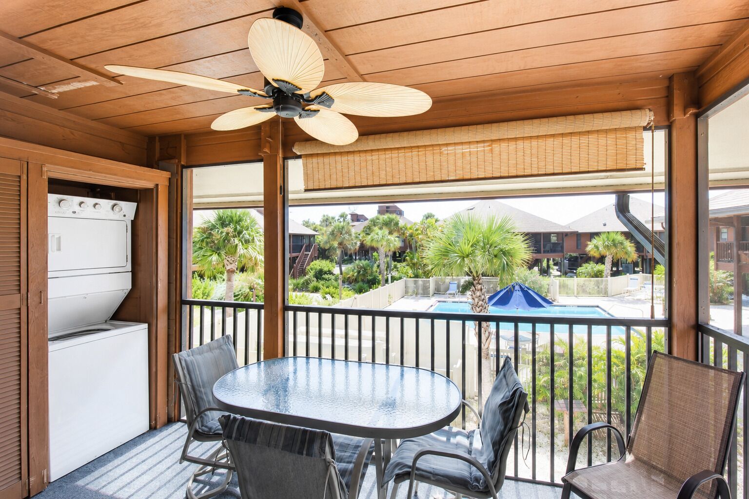 Un espace extérieur abrité, doté d’une table et de chaises, d’un ventilateur de plafond, et offrant une vue sur une piscine et des bâtiments en arrière-plan.