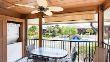 Un espace extérieur abrité, doté d’une table et de chaises, d’un ventilateur de plafond, et offrant une vue sur une piscine et des bâtiments en arrière-plan.