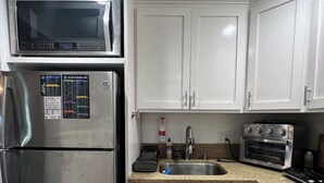 Cozinha compacta com armários brancos, geladeira de aço inoxidável, micro-ondas, forno elétrico e bancada de granito.