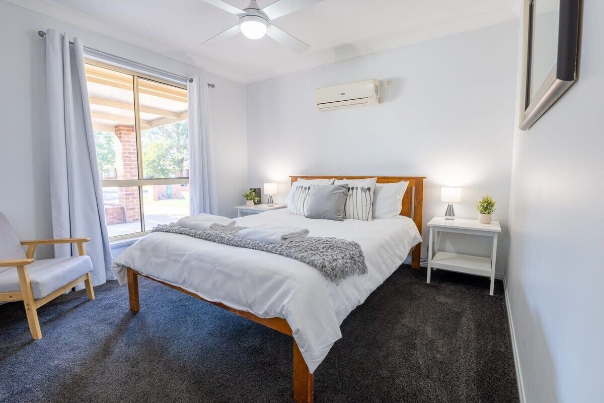 A bedroom with a bed, a chair, a bedside table, a nightstand, a ceiling fan, and an air conditioning unit.