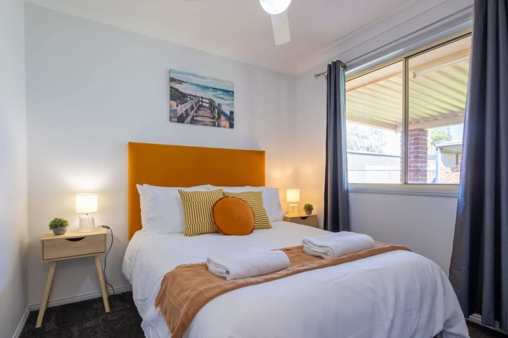 A hotel room with a bed, two bedside tables, a painting, a window with curtains, and a view of the outside.