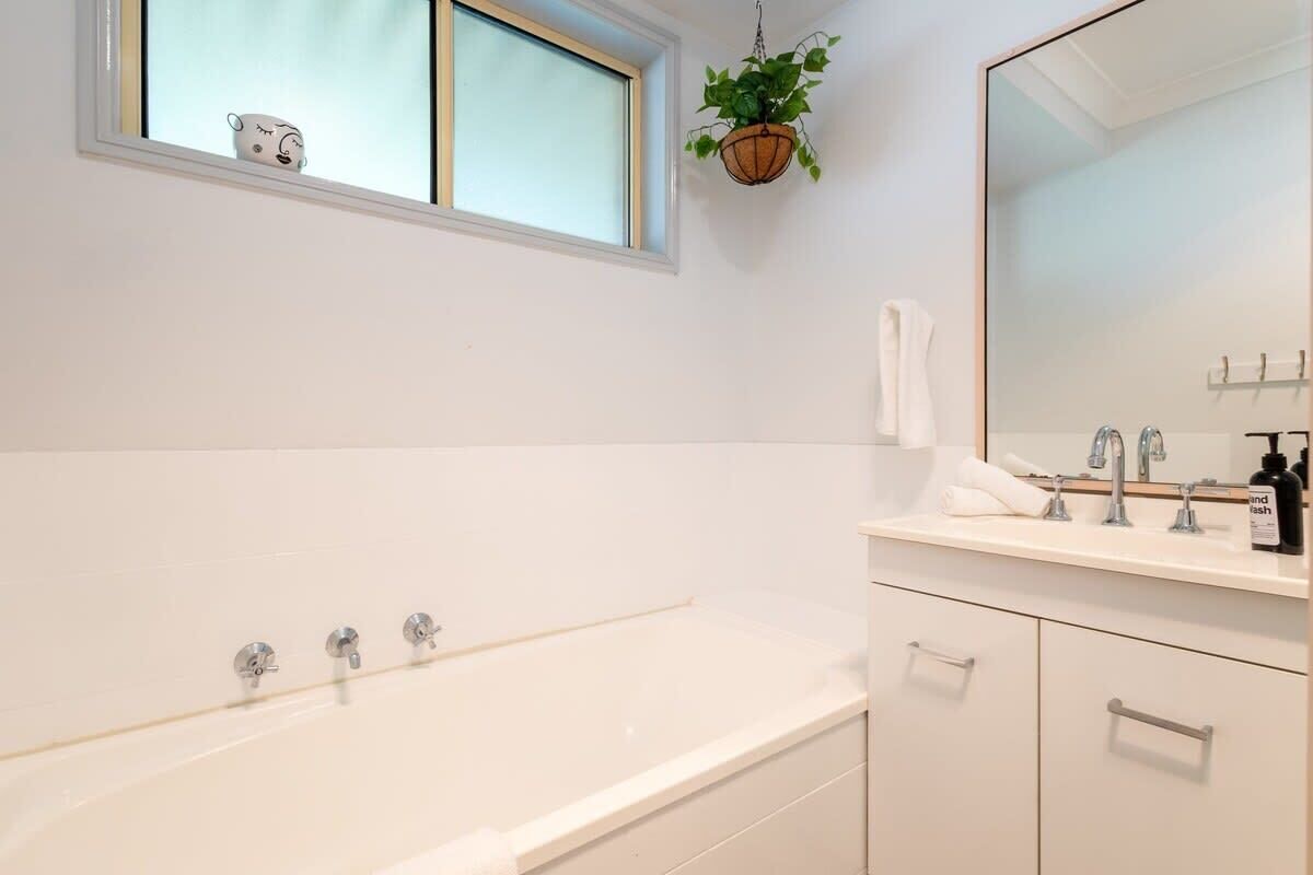 A modern bathroom with a large bathtub, a window with a plant, and a mirror above the sink.