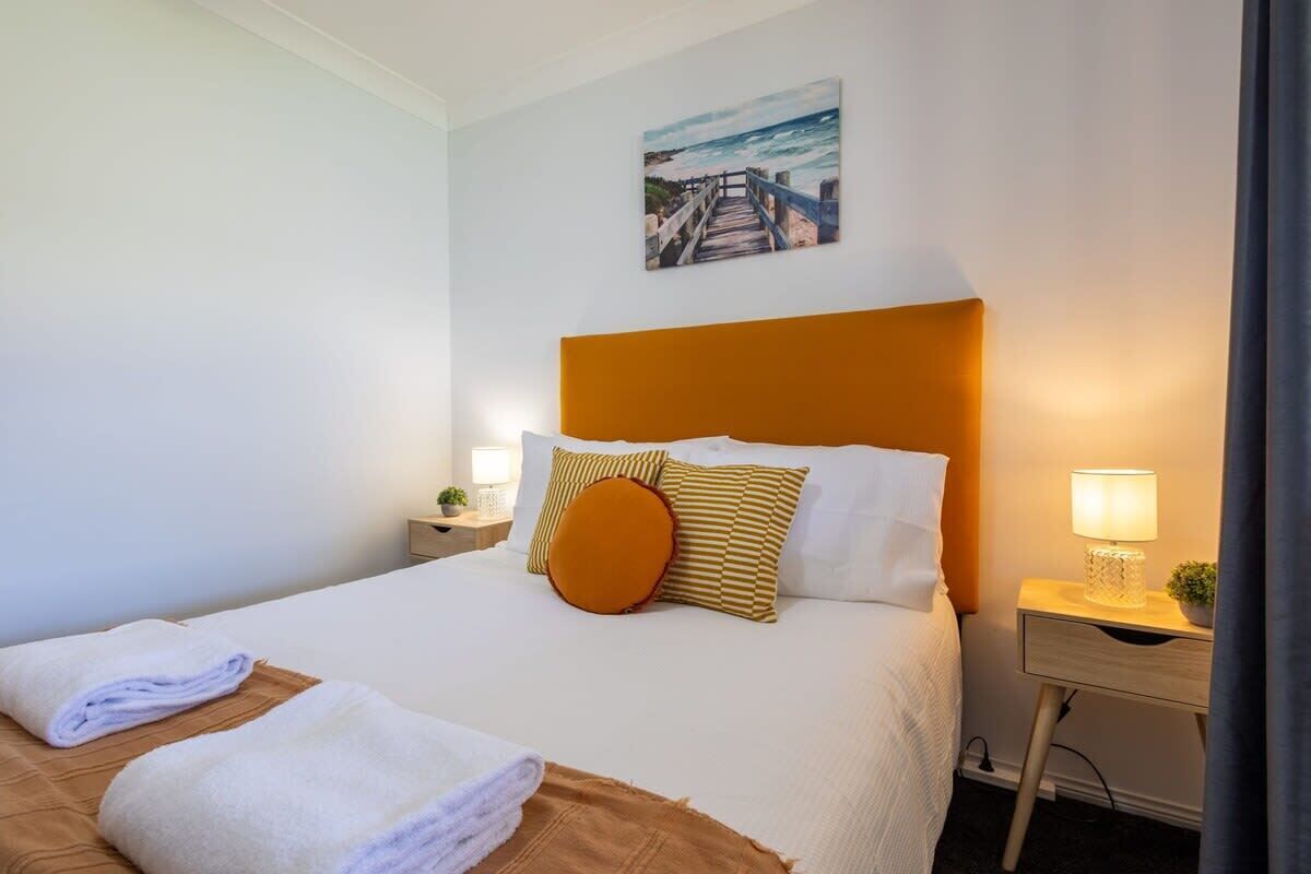 A neatly made bed with white linens and a brown headboard, a small bedside table with a lamp, and a painting of a beach scene above the bed.