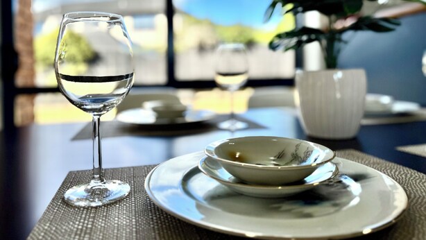 Een eettafel met een glas water, een kom en een bord, met uitzicht op de lucht en groen bij een raam.