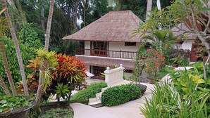 A two-story building with a thatched roof, surrounded by lush tropical vegetation and a paved pathway. - Kembali On The River by Balipras (Cepaka)