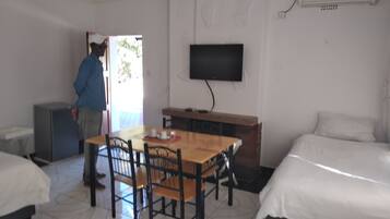 A hotel room with a wooden dining table, chairs, a TV mounted on the wall, a bed, and a small kitchenette area.