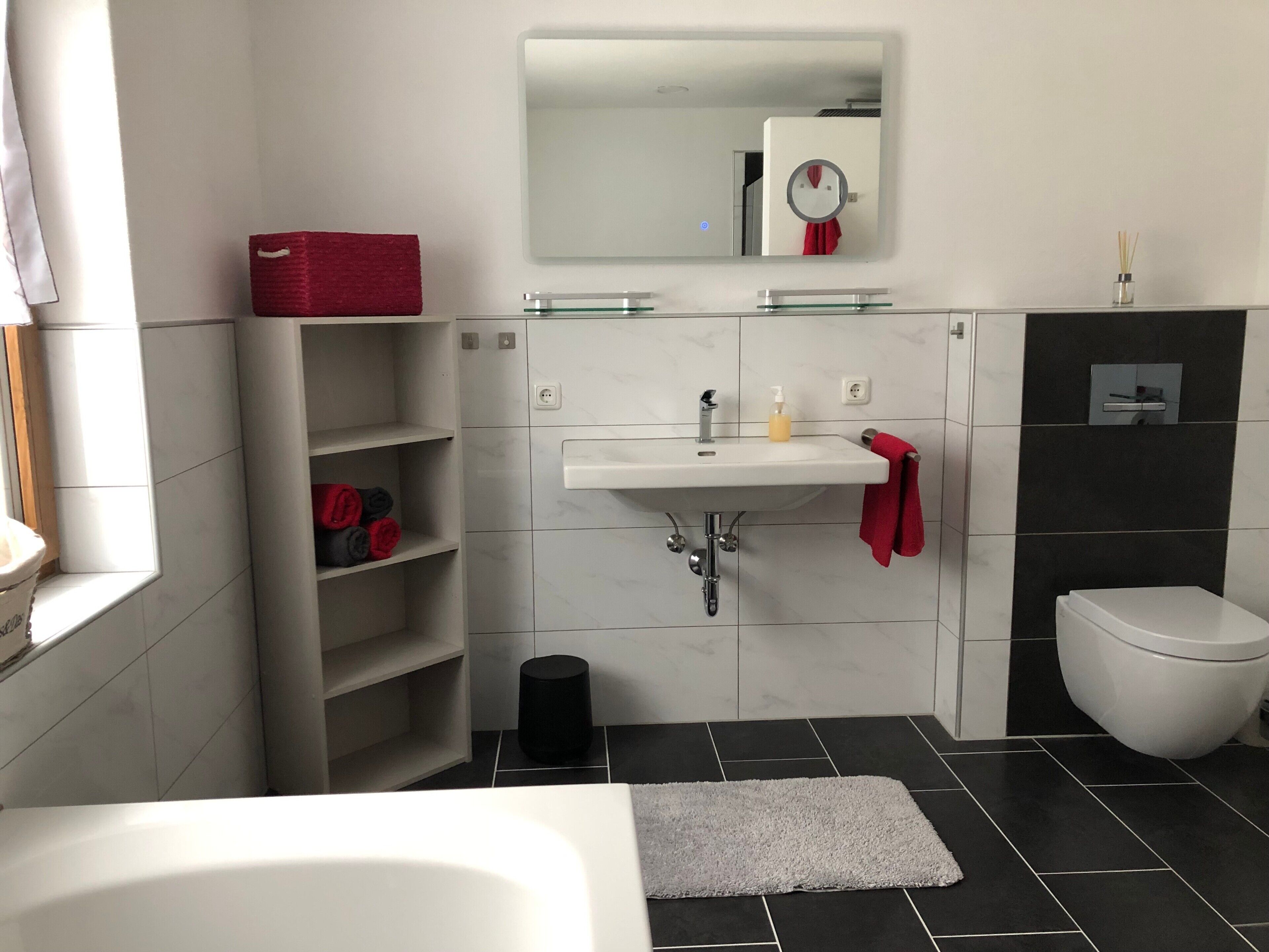 Un baño moderno con un lavamanos blanco, espejo y retrete. Hay estantes con toallas y un cubo de basura negro.