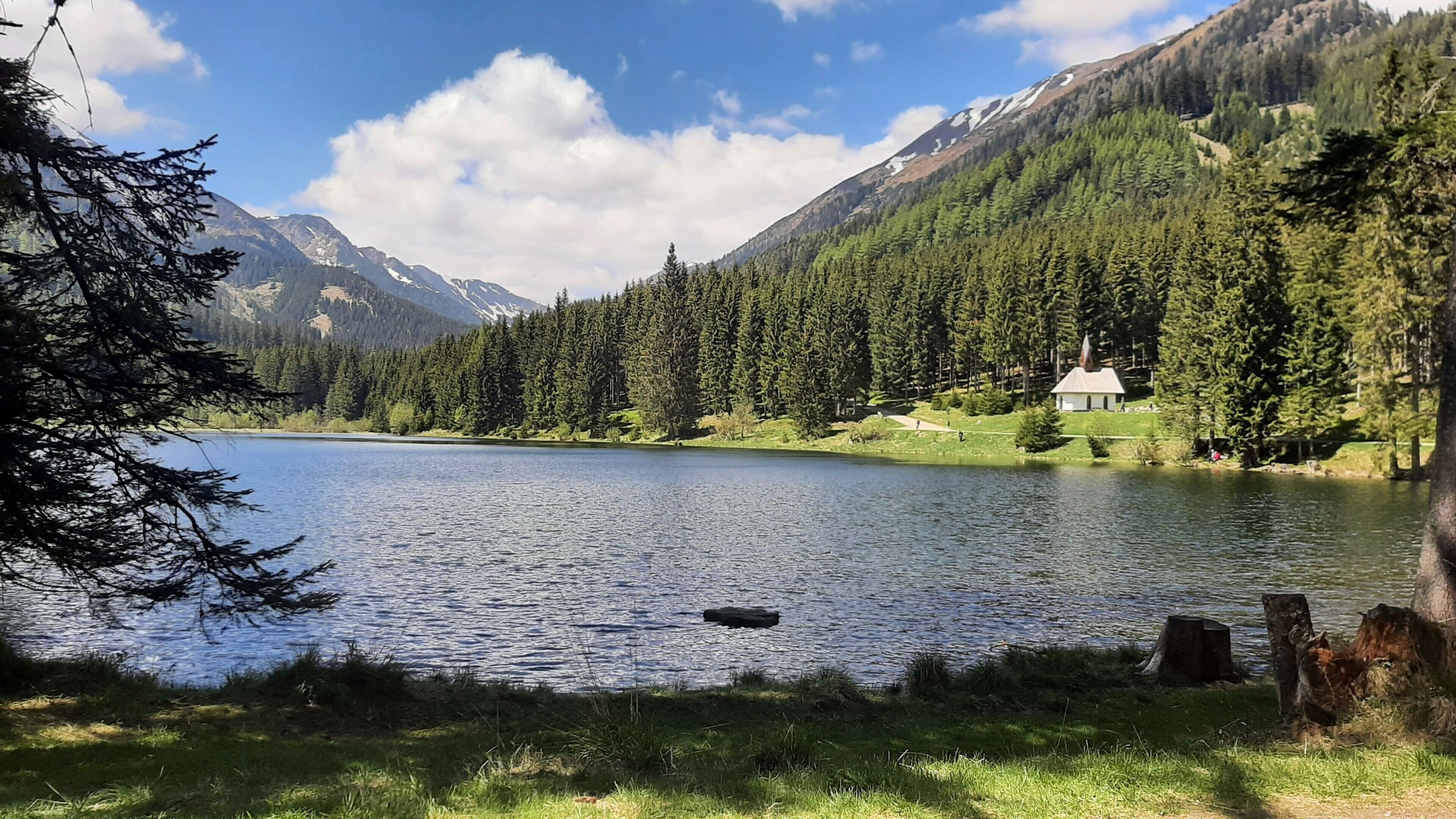 Un lago tranquilo con una cabaña pequeña y una iglesia al fondo, rodeado de un bosque frondoso y montañas.