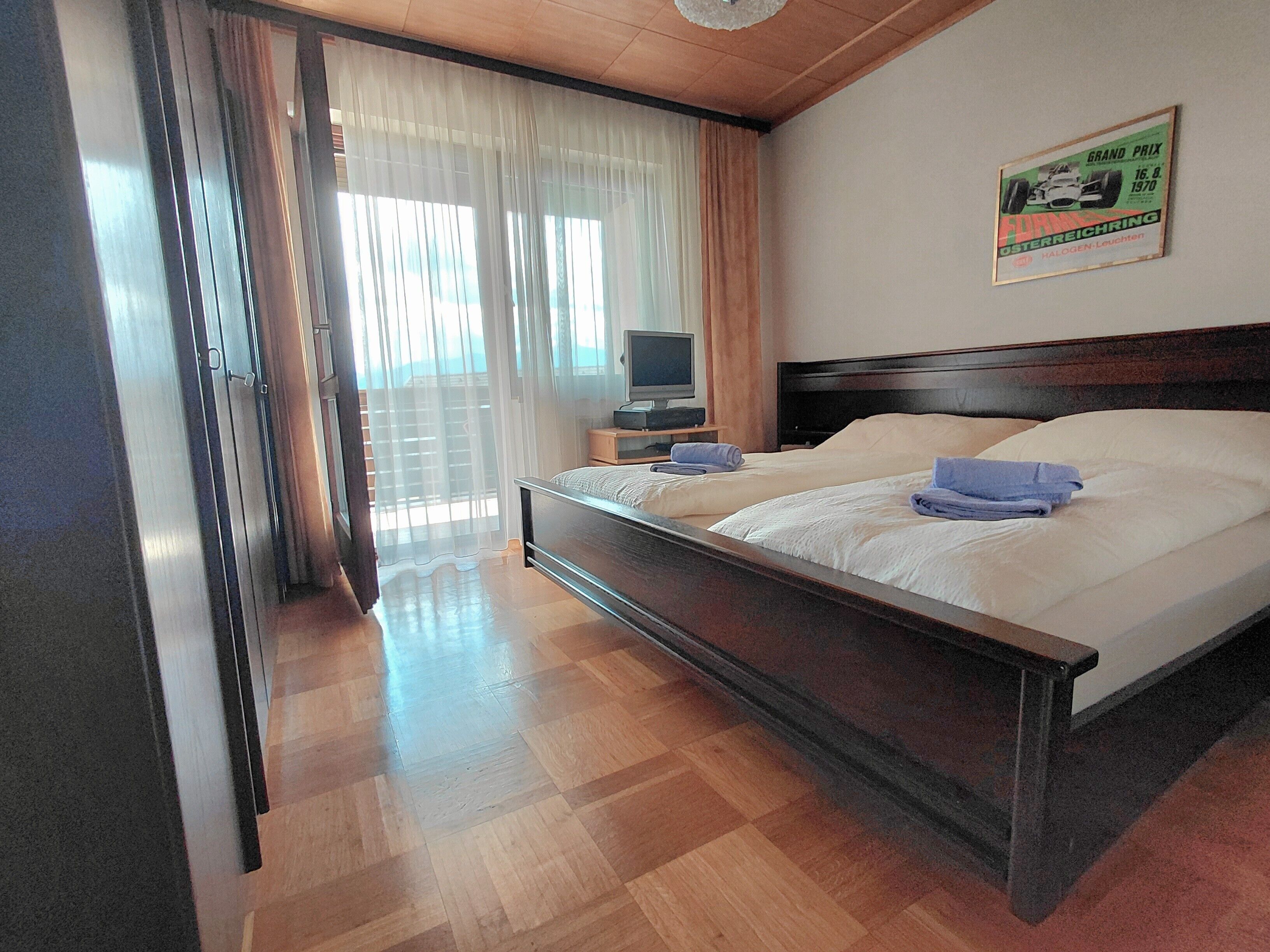 Une chambre d’hôtel avec un plancher en bois, un lit recouvert de draps blancs, un téléviseur posé sur une table de chevet et une fenêtre ornée de rideaux légers.