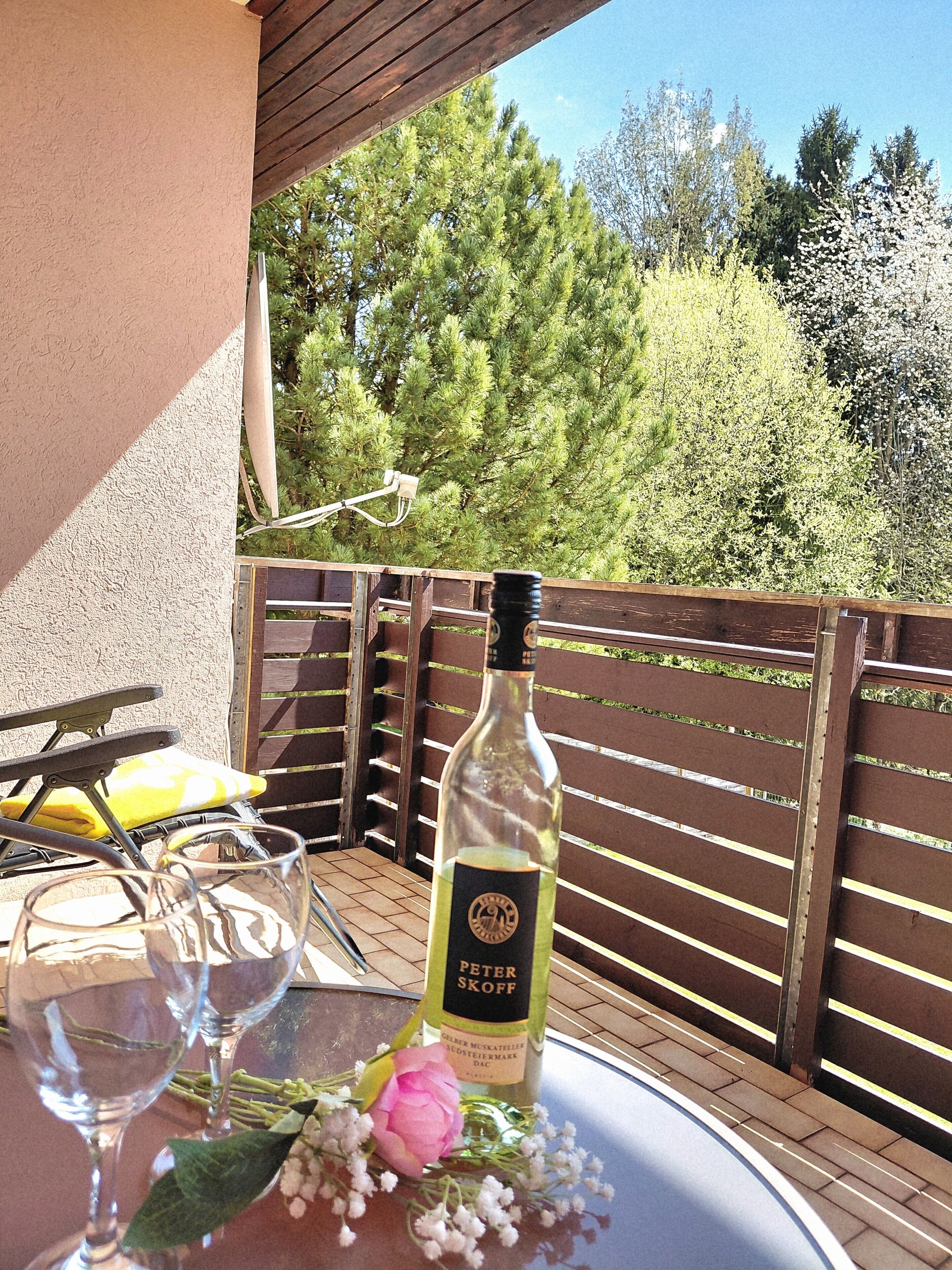 Ein Tisch mit einer Weinflasche, Gläsern und Blumen auf einer Terrasses mit Holzlattenzaun und Bäumen im Hintergrund.