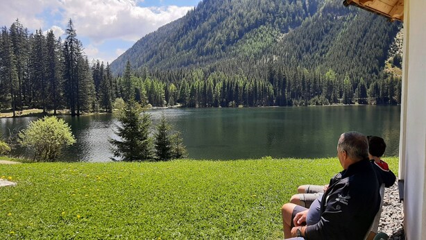 Une vue d’un lac entouré de montagnes boisées, avec une personne assise sur un banc.