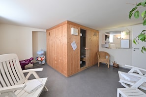A room with a wooden sauna, white chairs, a wicker chair, a small kitchen area, and a whiteboard.