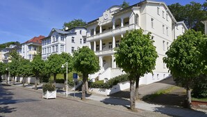 A row of white buildings with balconies, a paved street, and trees.