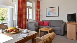 Un salon avec un canapé gris, une petite table en bois, un fauteuil en osier, une table à manger avec des fruits et une tasse, et un téléviseur posé sur un meuble.