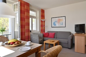 Una sala de estar con un sofá gris, una mesita de madera, una silla de mimbre, una mesa de comedor con fruta y una taza, y un televisor sobre un mueble.