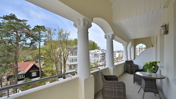 Un balcon agrémenté de meubles en osier, de colonnes blanches et offrant une vue sur les maisons.