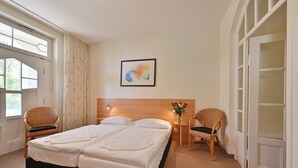 Ein Doppelbett mit Holz Kopfteil, zwei Nachttische mit Lampen, ein Korbstuhl und ein Fenster mit Vorhängen.