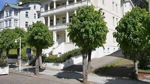 Ein großes, weißes Gebäude mit mehreren Stockwerken und einer zentralen Treppe. Vor dem Gebäude befinden sich Bäume, und ein Parkschild ist sichtbar.