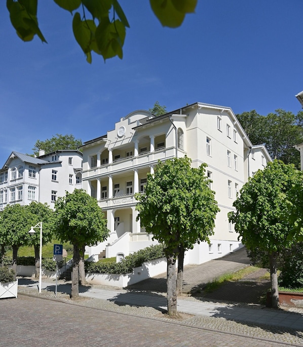 A large, white building with multiple floors and a central staircase. There are trees in front of the building, and a parking sign is visible.