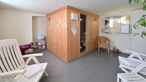 Une pièce comprenant un sauna en bois, des chaises blanches, un fauteuil en osier, une petite table et un tableau blanc.