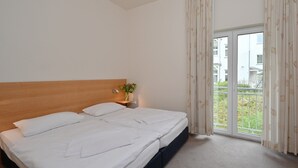 Un lit double avec une tête de lit en bois, une table de chevet avec une lampe, et une vue sur l’extérieur grâce à une porte-fenêtre coulissante qui donne sur un espace extérieur.