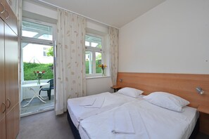A bedroom with a bed, a window with curtains, a small table with a chair, and a view of greenery outside.