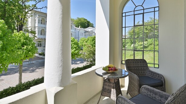 Un balcone con mobili in vimini, un tavolino con della frutta e vista su edifici e alberi.