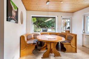 Un espace repas comprenant une table ronde en bois et des chaises rembourrées, de grandes fenêtres et une vue sur la verdure environnante.