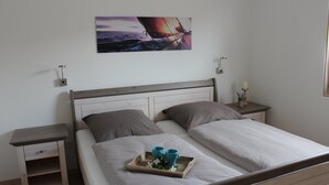 Un lit double avec une tête de lit en bois, deux tables de chevet, un tableau représentant un voilier accroché au mur, et un plateau avec deux tasses et des fleurs posé sur le lit.