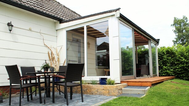 Une terrasse avec une table et des chaises, un espace couvert doté de portes-fenêtres coulissantes et un jardinière en pierre agrémentée de plantes.