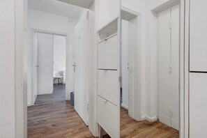 Un couloir aux murs blancs et au plancher en bois, qui mène à une pièce dotée d’une porte blanche.