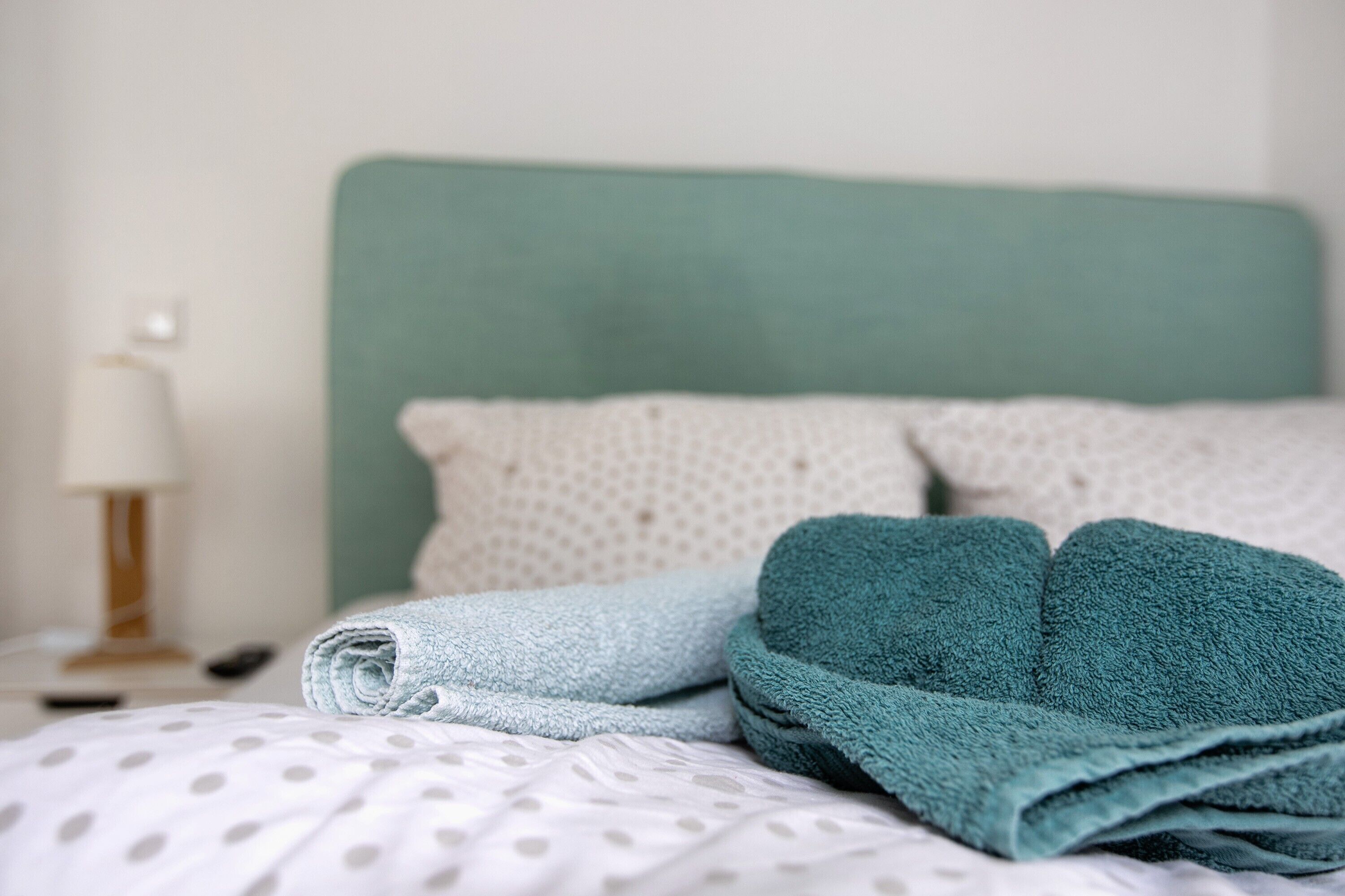 Una cama bien hecha con colcha de lunares, dos toallas enrolladas y un cabecero verde.