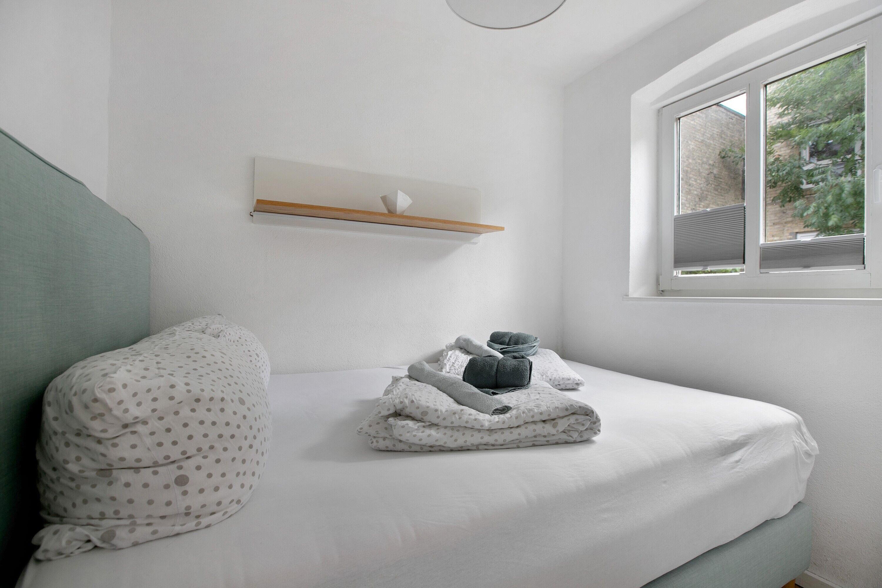 A neatly made bed with folded towels and a pillow, a small shelf above it, and a window with a view of greenery.