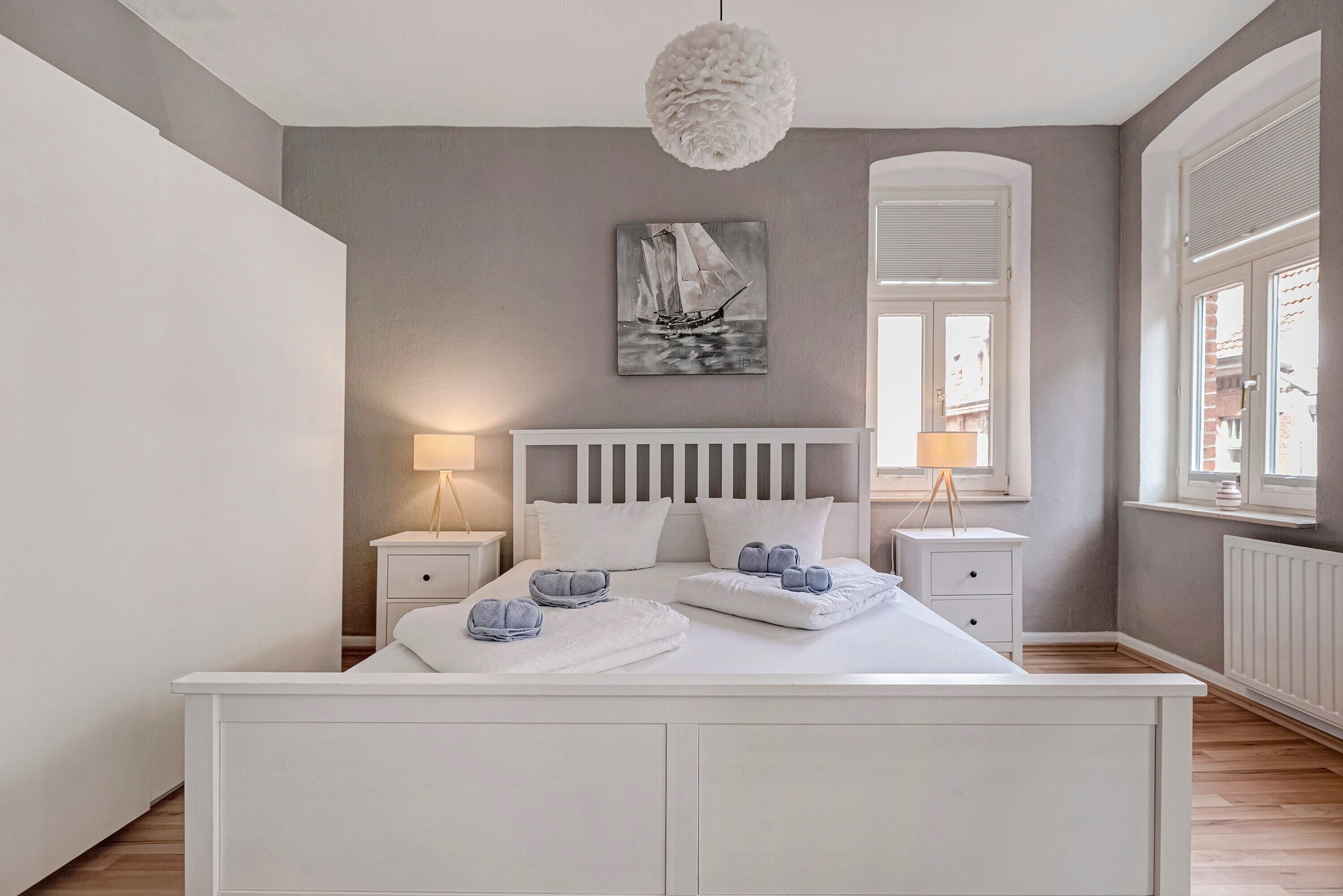Un dormitorio con una cama blanca, dos mesitas de noche, una lámpara colgante, una ventana con persianas y un cuadro en la pared.
