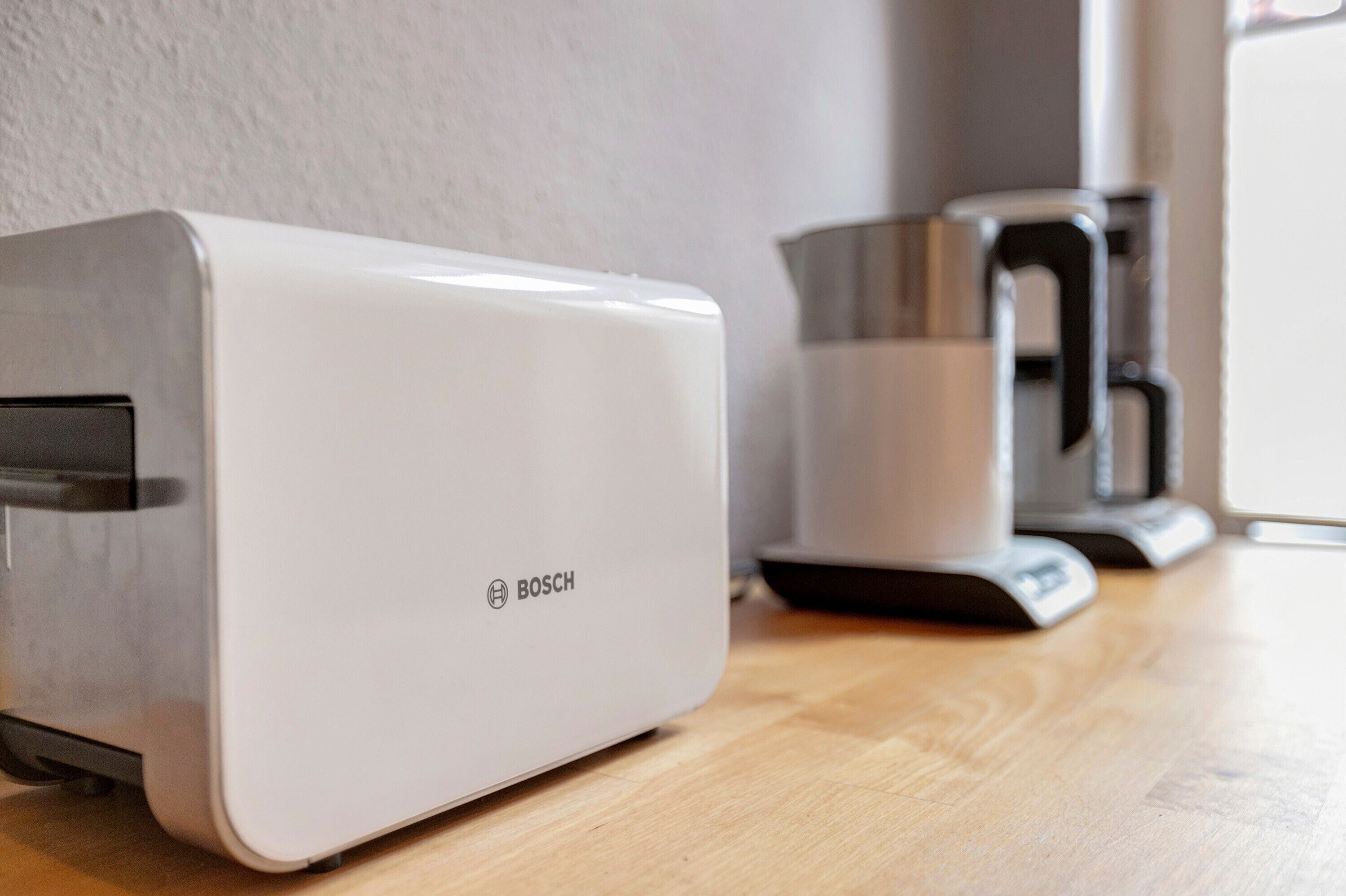 Un tostador blanco Bosch sobre una mesa de madera, junto a una cafetera de acero inoxidable.