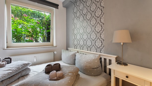 Een slaapkamer met een raam, een radiator, een bed met kussens, een nachtkastje en een lamp.