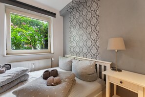 A bedroom with a window, a radiator, a bed with pillows, a bedside table, and a lamp.