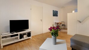 Een woonkamer met een flatscreen televisie op een witte zuil, een kleine witte tafel met een bloemenvaas, een grijze bank en een eethoek met stoelen en een tafel op de achtergrond.
