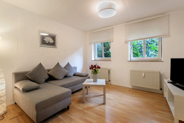 A modern living room with a grey sofa, a small white table with a vase of flowers, a television on a stand, and large windows with blinds.